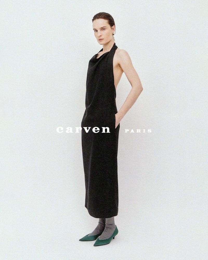 Charlotte Tomas in a black halter backless dress, grey socks, and green kitten heels against a white studio background for Carven.