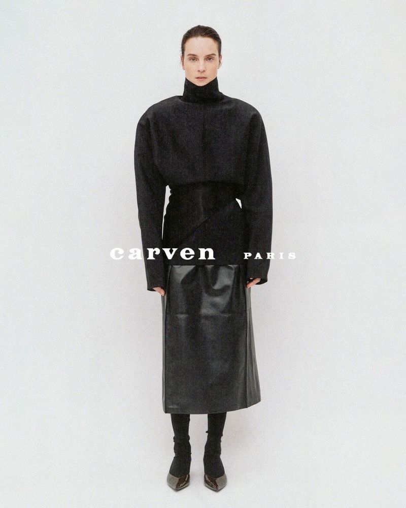 Model Charlotte Tomas wearing a structured high-neck black top and leather midi skirt against a plain white studio backdrop.