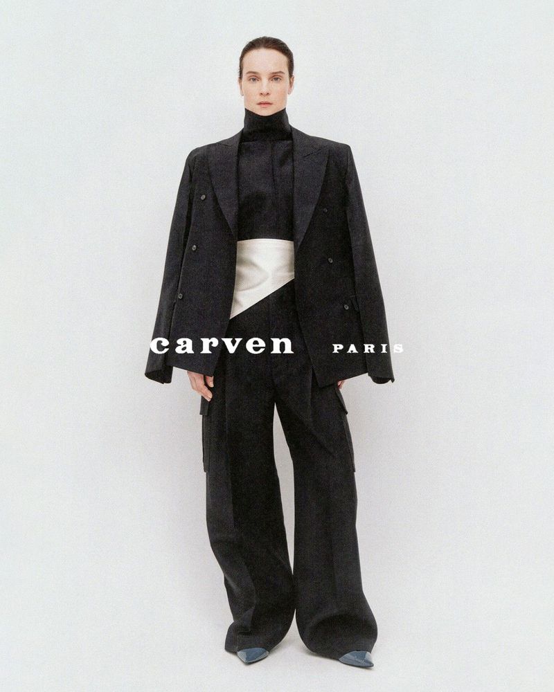 Model Charlotte Tomas in an oversized black suit with draped blazer and white sash against a studio background.