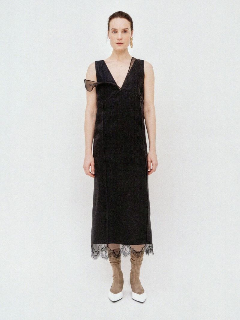 Model Charlotte Tomas poses in a black sleeveless V-neck midi dress with lace hem trim and white pointed heels on a white backdrop.