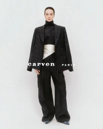 Model Charlotte Tomas in an oversized black suit with draped blazer and white sash against a studio background.
