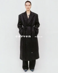 Model in a long black belted coat and trousers against a white seamless background with Carven Paris branding.