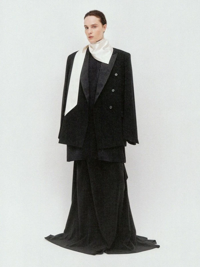 Charlotte Tomas posing in an oversized black tailored jacket, long draped skirt, and off-white scarf against a seamless white background.