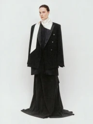 Charlotte Tomas posing in an oversized black tailored jacket, long draped skirt, and off-white scarf against a seamless white background.
