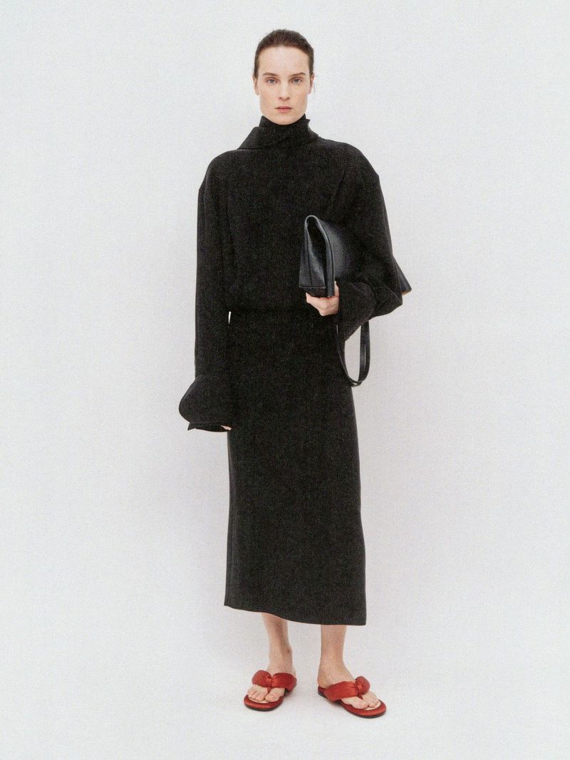 Model Charlotte Tomas wearing a black turtleneck midi dress with a black clutch and red sandals against a white background.