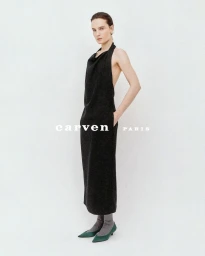 Charlotte Tomas in a black halter backless dress, grey socks, and green kitten heels against a white studio background for Carven.