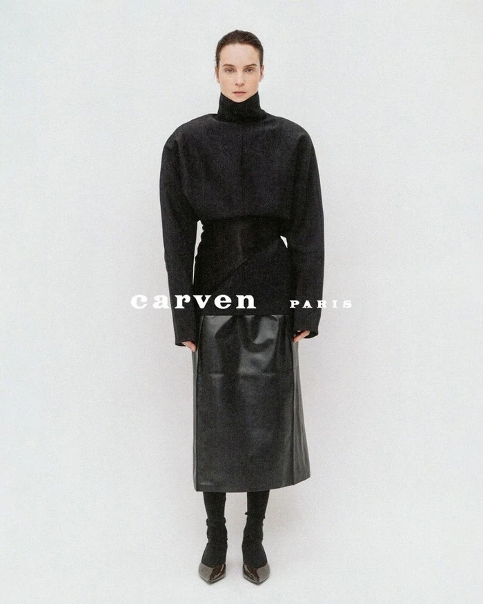 Model Charlotte Tomas wearing a structured high-neck black top and leather midi skirt against a plain white studio backdrop.
