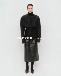 Model Charlotte Tomas wearing a structured high-neck black top and leather midi skirt against a plain white studio backdrop.