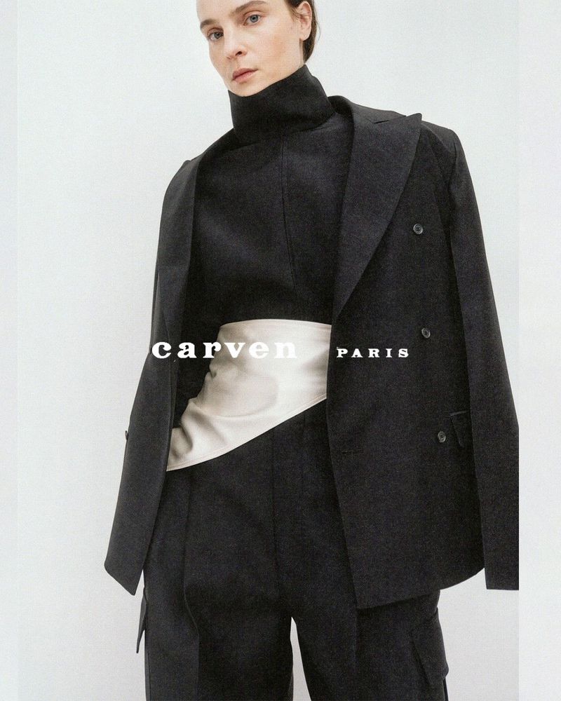 Model wearing a black tailored blazer, turtleneck top, and an ivory satin waist sash against a neutral backdrop.