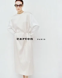 Model Charlotte Tomas wearing black glasses and a belted off-white dress against a white studio background for Carven.