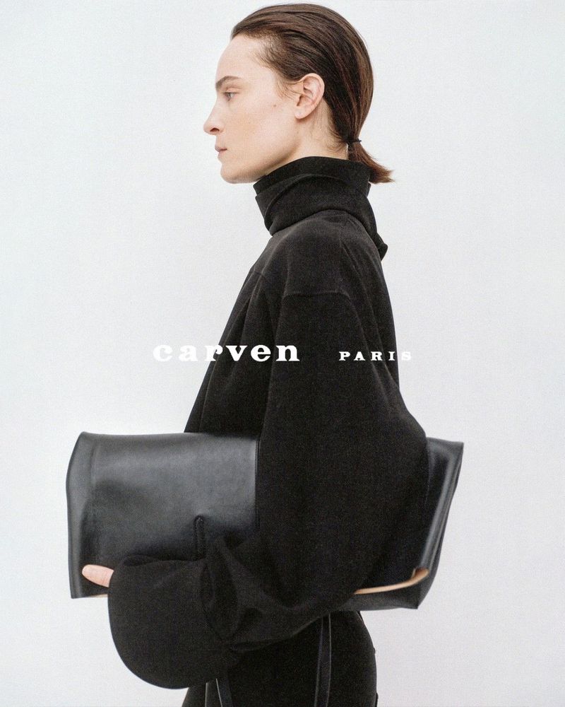 Model Charlotte Tomas in profile holding an oversized black leather clutch bag and wearing a black turtleneck against a white backdrop.
