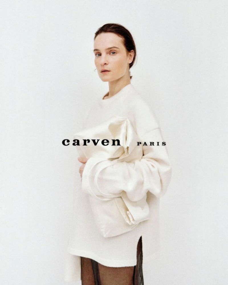 Model Charlotte Tomas holding a soft white bag while wearing an oversized cream sweater against a white background.
