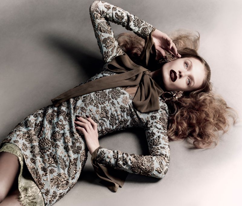Model Ella McCutcheon lying down in a gold-embroidered dress with a large neck bow against a grey backdrop.