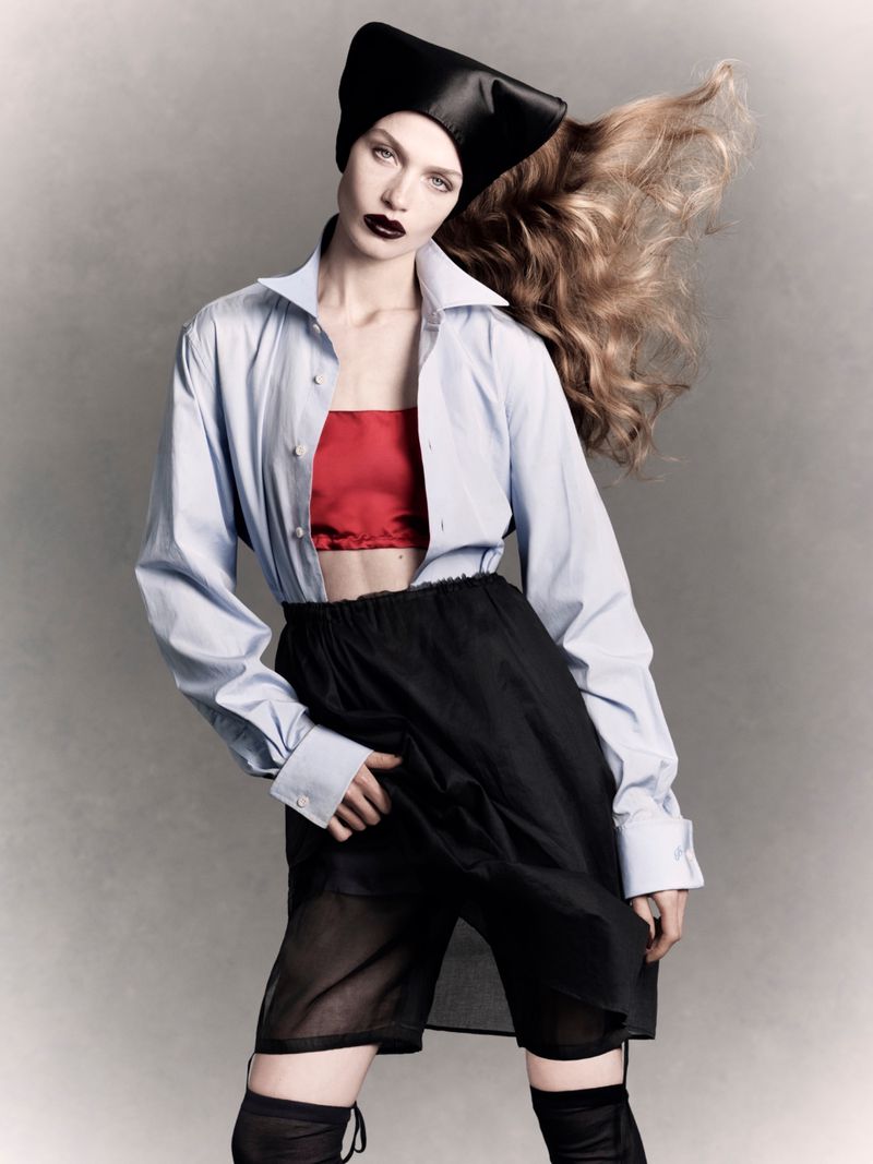 Model Ella McCutcheon posing in a light blue open shirt, red bandeau, black sheer skirt, and black headpiece against a grey backdrop.