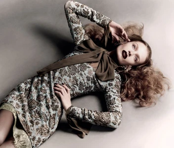Model Ella McCutcheon lying down in a gold-embroidered dress with a large neck bow against a grey backdrop.