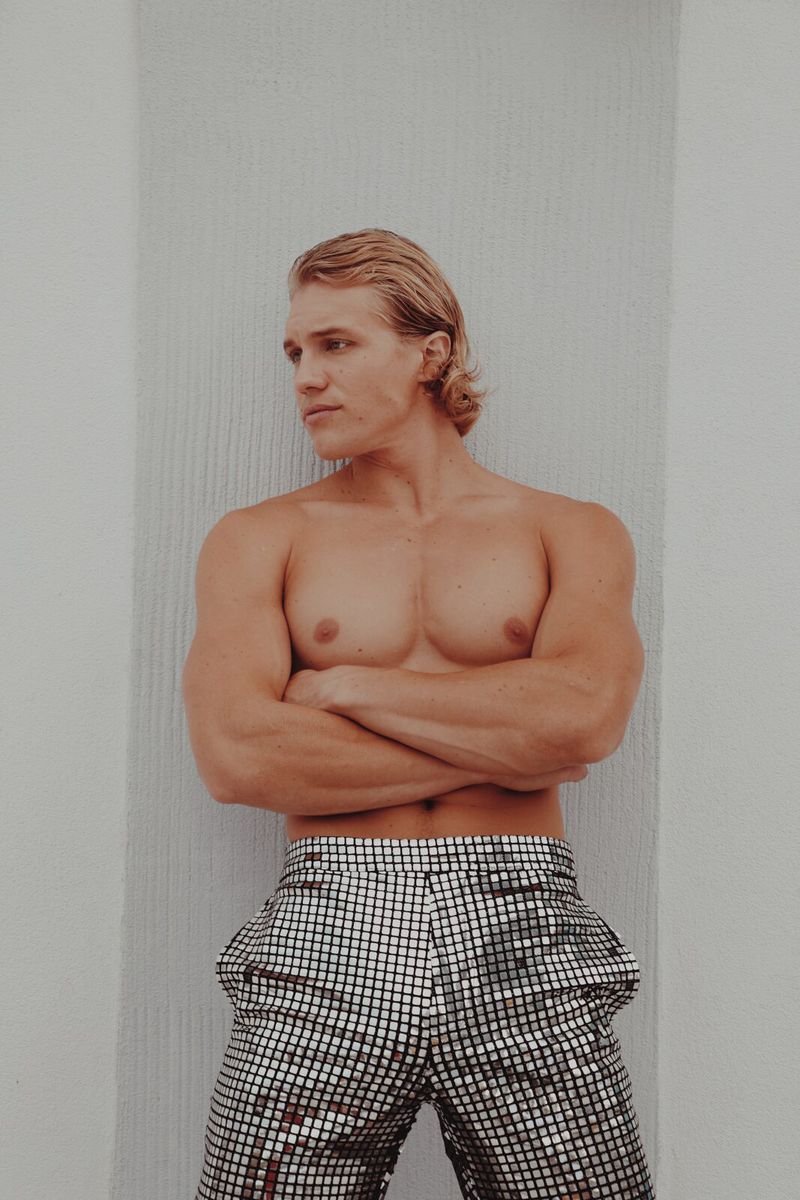 Blond male model in silver disco pants poses with arms crossed against a white wall. Fashion shot in bright light.