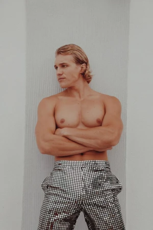 Blond male model in silver disco pants poses with arms crossed against a white wall. Fashion shot in bright light.