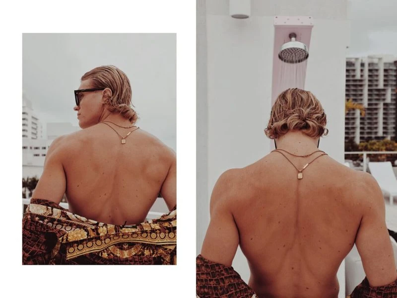 Muscular male model with blond hair poses outdoors with patterned robe, facing away from the camera, with a city skyline in the background.