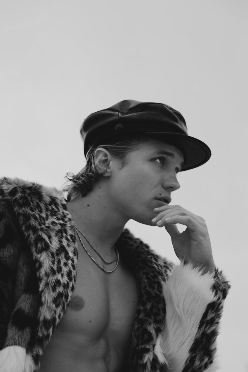 Monochrome portrait of a young adult male wearing a faux fur coat and cap, hand to face, looking up against a neutral background.