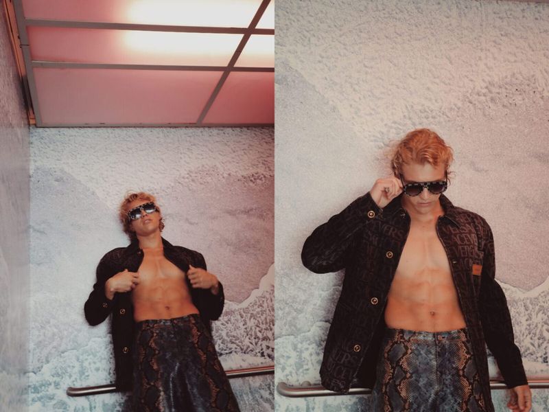 A blonde male model poses shirtless in a textured jacket and printed pants, indoors against a white wall with overhead lighting.
