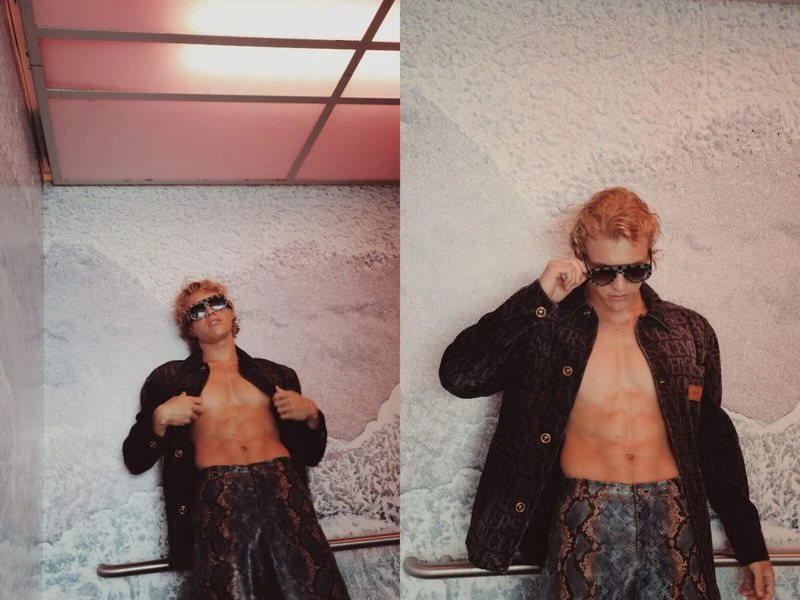 A blonde male model poses shirtless in a textured jacket and printed pants, indoors against a white wall with overhead lighting.