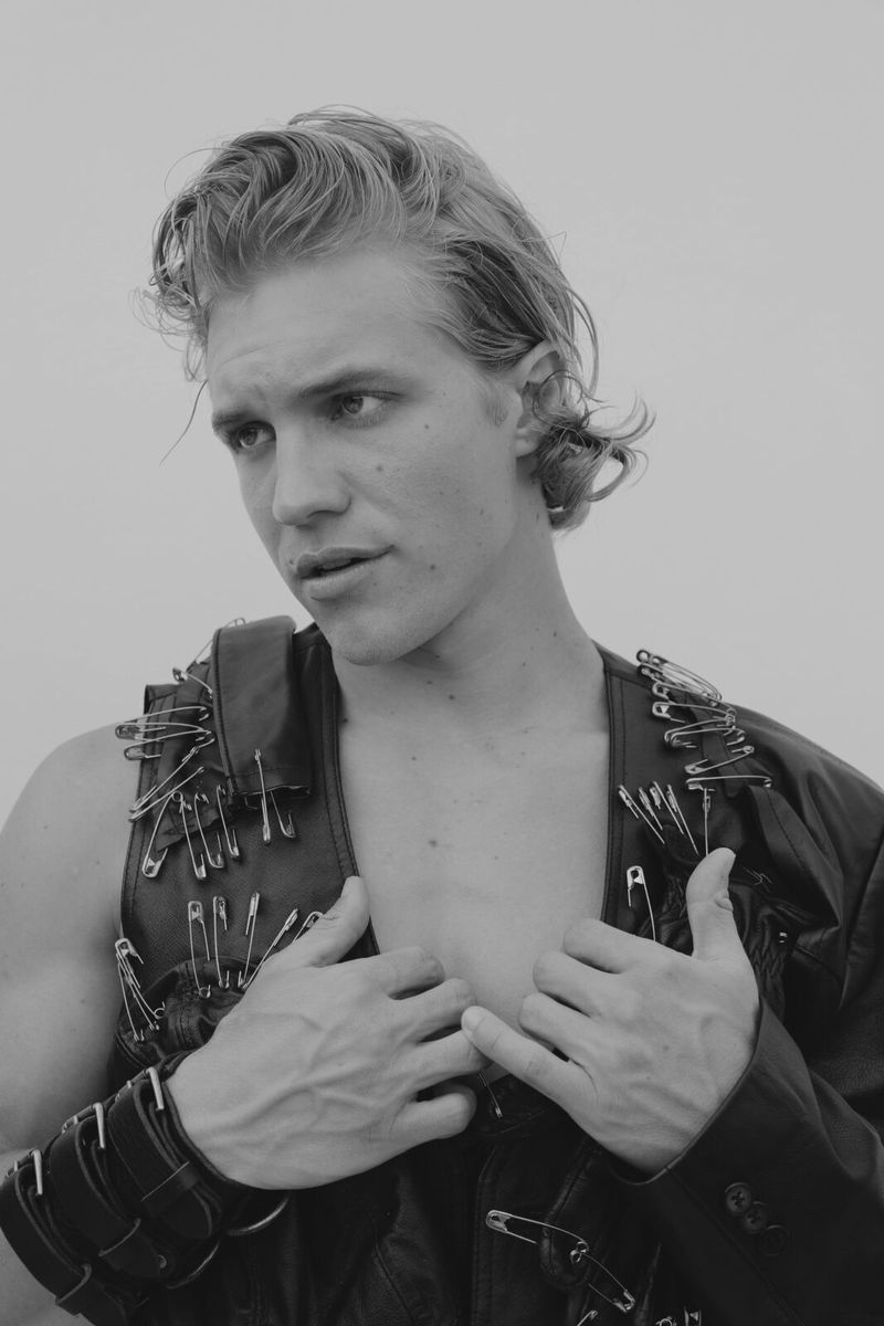 Black-and-white portrait of a fair-skinned male adult wearing a safety pin-studded leather vest with a jacket and metal bracelets.