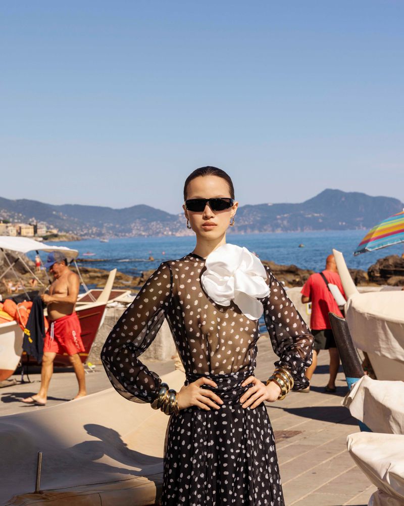 Model wearing a black sheer polka dot dress, white flower collar, and sunglasses standing by a coastal seaside harbor