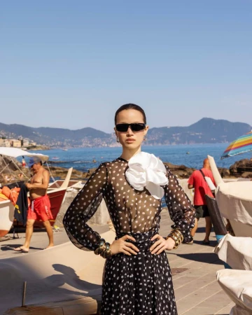 Model wearing a black sheer polka dot dress, white flower collar, and sunglasses standing by a coastal seaside harbor