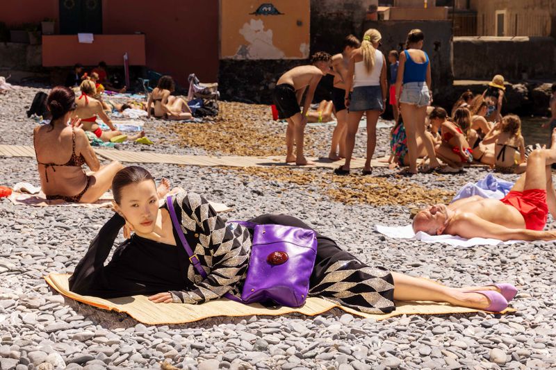 Model lying on a pebble beach wearing a diamond pattern cardigan with a purple leather bag among sunbathers in bright sunlight.