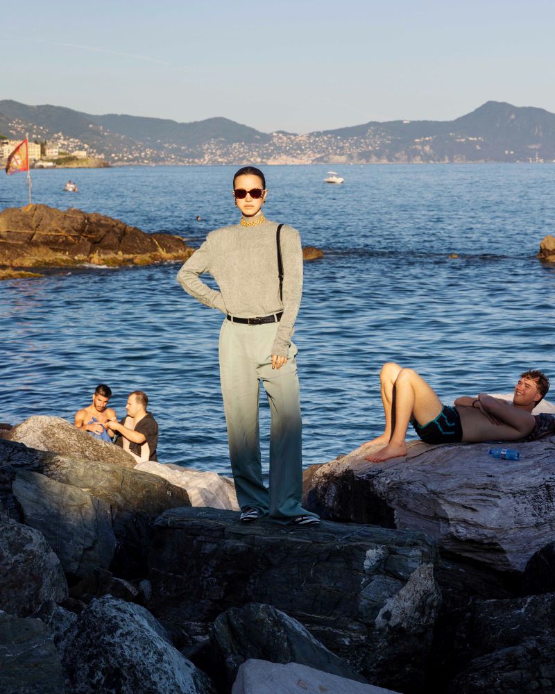 Model wearing sunglasses and grey trousers standing on rocks by the sea, with other people relaxing in the background