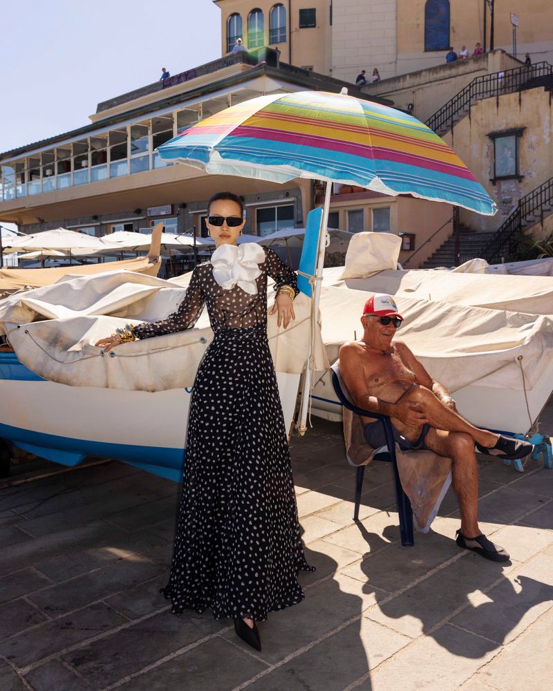 A woman in a polka dot dress standing beneath a striped beach umbrella next to boats, with a seated man nearby.
