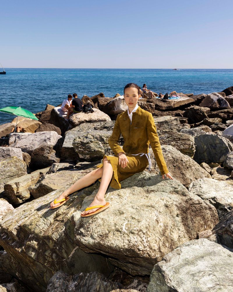 An East Asian woman wearing an olive tailored suit and yellow flip-flops sits on large coastal rocks overlooking the blue sea.