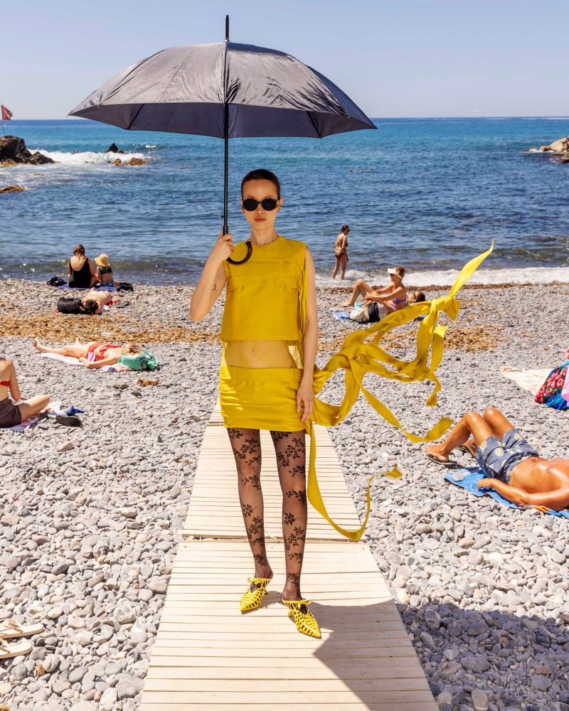 Model standing on a wooden beach path holding a black umbrella wearing a yellow two-piece set, patterned tights, and yellow mules.