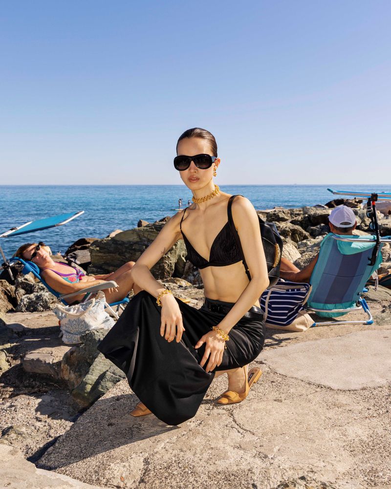 Woman in black bikini top and skirt wearing oversized sunglasses posing on a rocky beach with ocean background