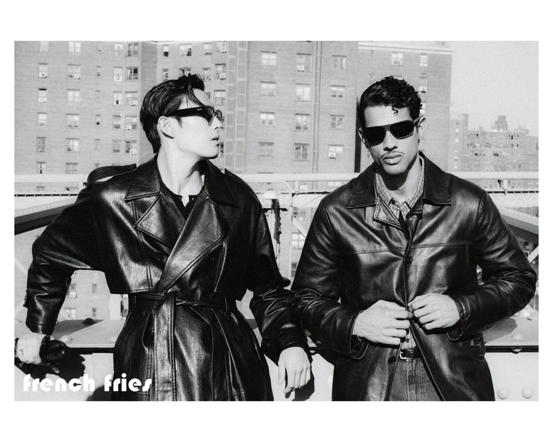Two men wearing black sunglasses and leather jackets pose along an outdoor bridge railing against city buildings.