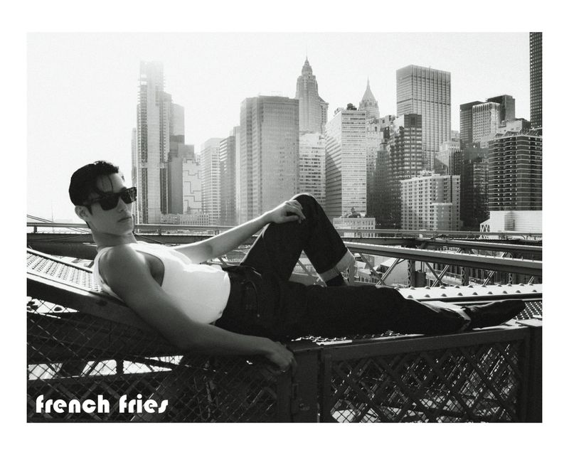 East Asian man in dark sunglasses and a white tank top reclining on a steel bridge structure with a city skyline behind him.