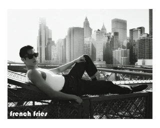 East Asian man in dark sunglasses and a white tank top reclining on a steel bridge structure with a city skyline behind him.
