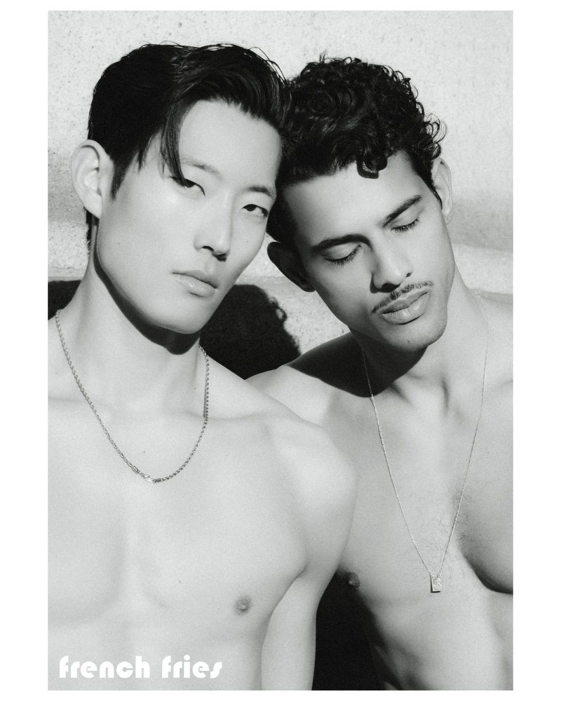 Black and white editorial portrait of two shirtless male models wearing chain necklaces against a concrete wall.