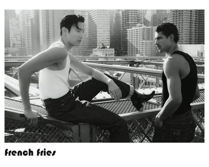Two male models in tank tops and jeans pose on a metal bridge structure overlooking city skyscrapers in black and white.