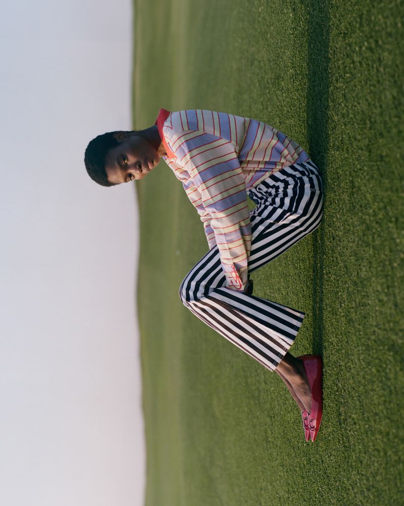 Fashion portrait of African-American woman in striped pants and shirt, leaning on a grassy hill in sunlight.