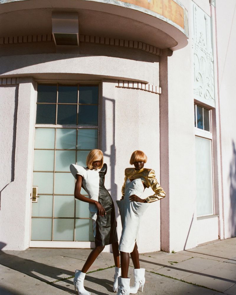Two Black women in designer dresses and white boots pose against an art deco building on a sunny day; fashion, style, urban setting.