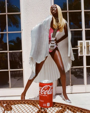 A tall, fashionable Black female adult poses outside in front of a closed door with a can of Coca-Cola at her feet in daylight.