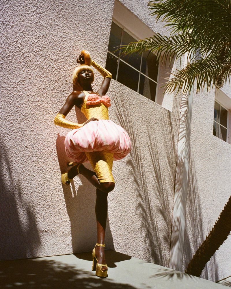 Woman in gold dress and pink skirt poses with palm trees and white building background in outdoor fashion shot, showcasing textures and shadows.