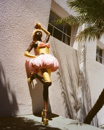 Woman in gold dress and pink skirt poses with palm trees and white building background in outdoor fashion shot, showcasing textures and shadows.