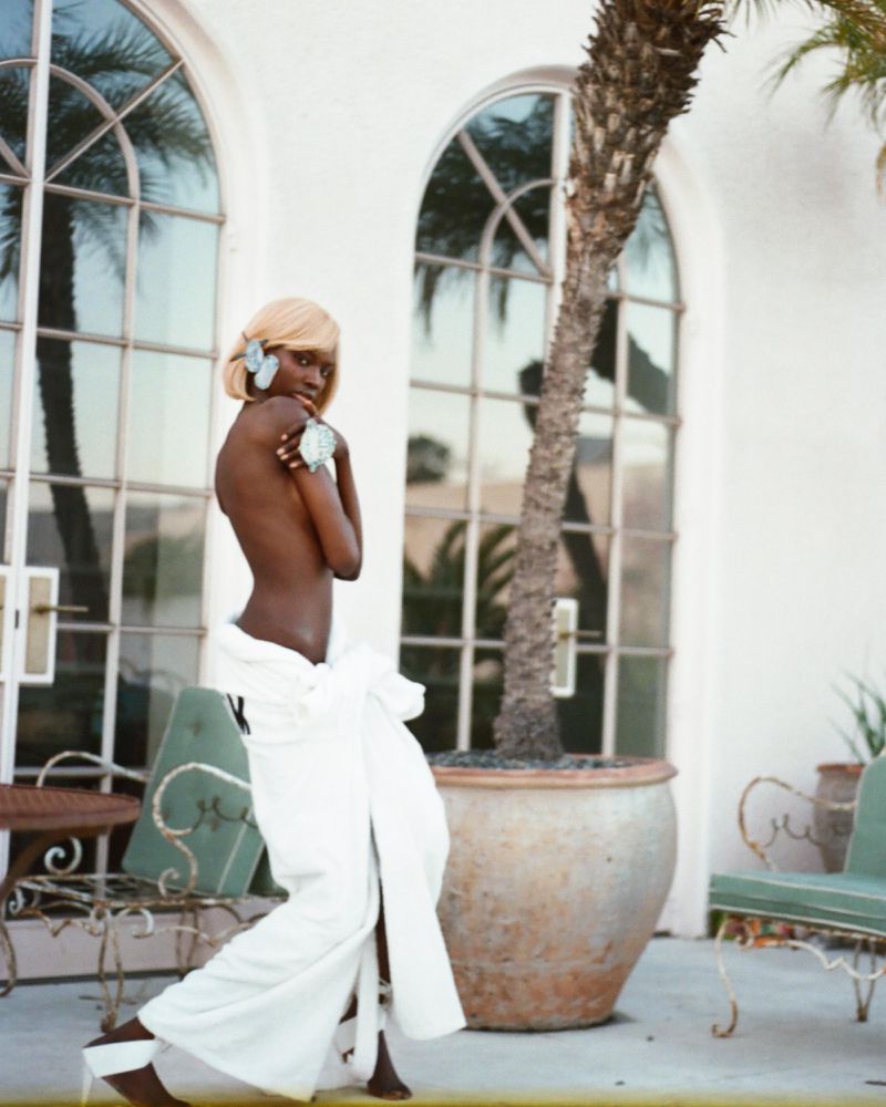 Elegant woman with blonde wig and white fabric wrap poses near a potted palm tree and white building with arched windows outside.