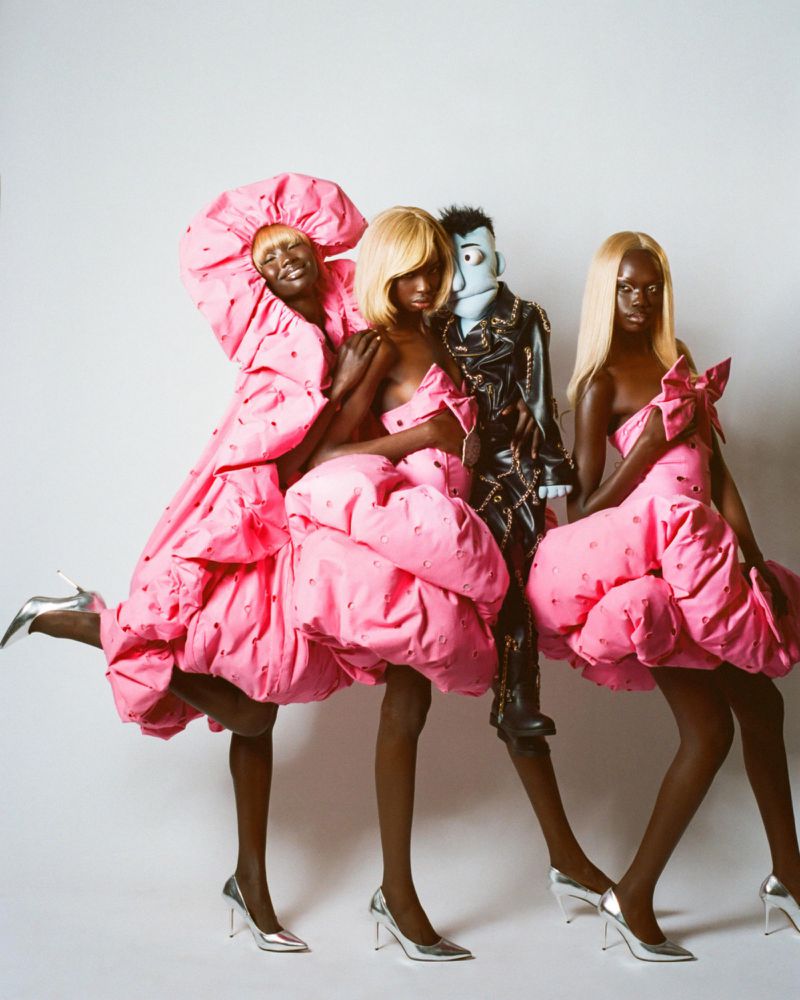 Three models and a puppet pose in pink dresses and silver heels against a white backdrop in a studio fashion editorial shot.