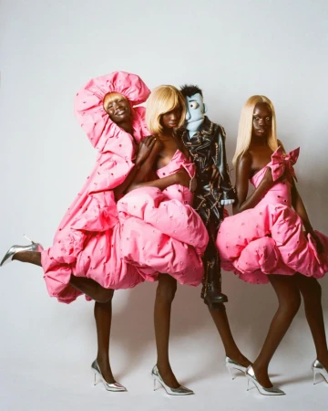 Three models and a puppet pose in pink dresses and silver heels against a white backdrop in a studio fashion editorial shot.