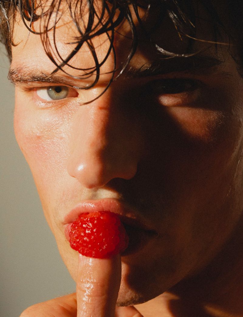Close-up portrait of model Stefano Tomadini holding a red raspberry on his finger to his lips under warm directional lighting.