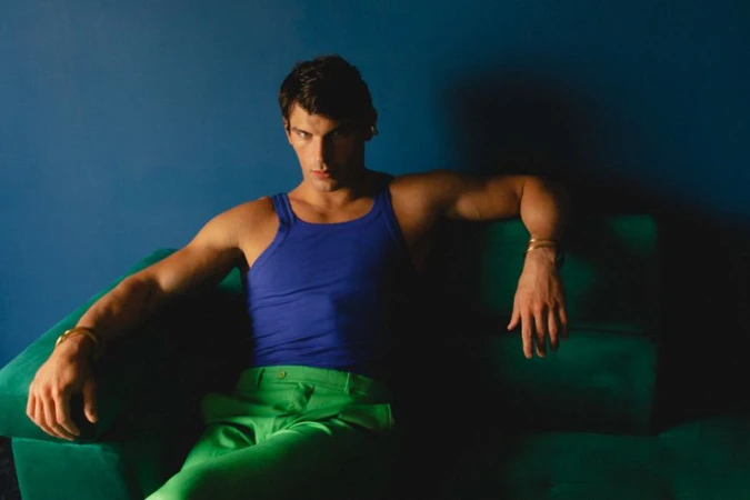 Model Stefano Tomadini seated on a green couch wearing a blue tank top and green pants against a dark blue wall.