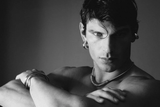 Black and white portrait of model Stefano Tomadini shirtless with arms crossed, wearing silver earrings, necklace, and bangles.